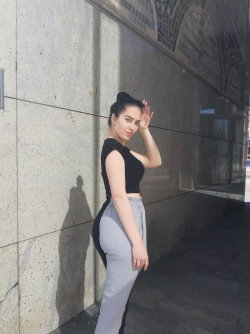 A woman with dark hair in a bun wears a black crop top and gray pants, leaning against a stone wall outdoors.