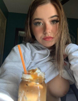 A woman with long brown hair and makeup wears a gray hoodie, holding a drink in a mason jar; one breast is exposed.