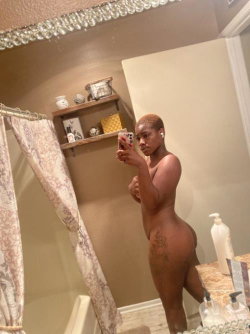 A Black woman with short hair takes a mirror selfie in a bathroom, standing nude with visible tattoos on her thigh.