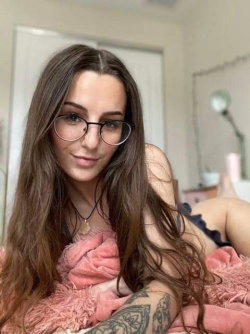 A woman with long brown hair and glasses lies on a pink blanket, wearing a dark top with ruffled edges; a tattoo is
