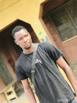 A young Black man with short hair and a beard wears a black t-shirt and a crossbody bag, standing outside a building