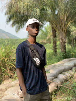 Young Black man in a navy t-shirt and white cap stands outdoors near palm trees, wearing a black Nike crossbody bag.