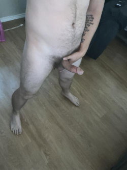 Shirtless man with visible pubic hair and arm tattoo, standing barefoot on wooden floor, holding his erect penis.