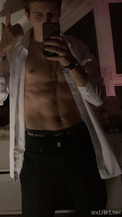 A slim, shirtless man in a white open shirt and black pants takes a mirror selfie, showing defined abs and wearing a