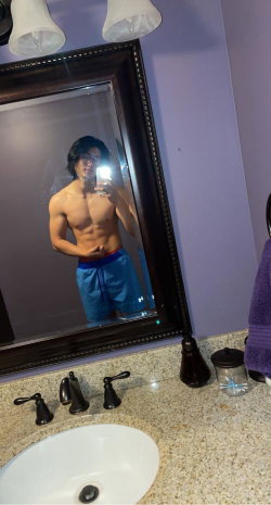 Shirtless muscular man with dark hair takes mirror selfie in bathroom, wearing blue shorts with red waistband.