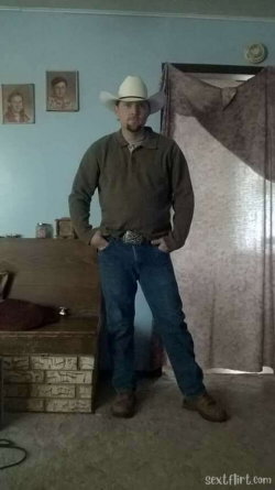 Man in cowboy hat and jeans stands confidently in a room with framed photos on the wall.