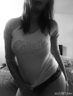 A woman with dark hair in a tight long-sleeve shirt and lace underwear poses on a bed, lifting her shirt slightly.