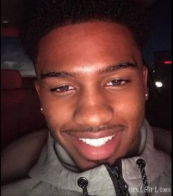 A smiling young Black man with short curly hair, wearing a gray hoodie, taking a selfie in a dimly lit car interior.