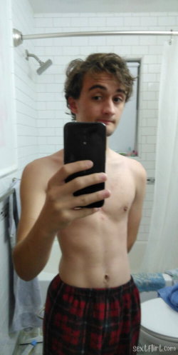 Shirtless man with curly hair taking a mirror selfie in a bathroom, wearing red plaid shorts.