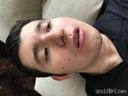 A young man with dark hair and stubble lies on a couch, wearing a black shirt, looking directly at the camera.