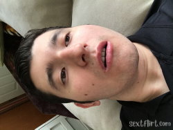 A young man with dark hair and stubble lies on a beige couch, looking at the camera in a close-up selfie.