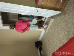A woman in a bright pink shirt and light shorts stands in a doorway, smiling at the camera with hands on hips.