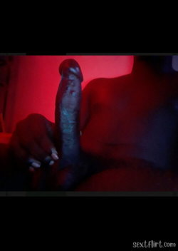 A person with dark skin holds a large erect penis under red lighting, partially visible torso and hand in frame.