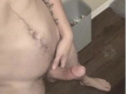 Close-up of a person's lower torso and legs, showing pubic hair, a tattooed arm, and bare feet on wooden flooring.