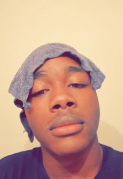 A young Black man with a towel on his head, wearing a dark blue shirt, looking directly at the camera in a plain indoor