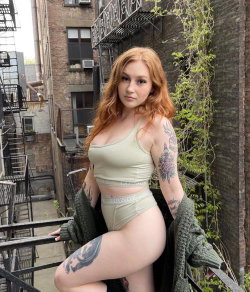 Redheaded woman with tattoos poses in a beige crop top and matching underwear on an urban balcony, holding a green