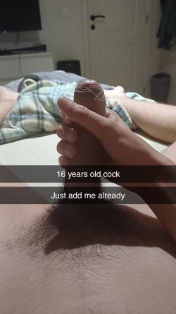 Close-up of a hand holding an erect penis, with legs and plaid shorts visible in the background on a bed.