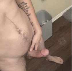 A shirtless person with visible chest hair and a tattooed arm is holding their erect penis, standing on a wooden floor