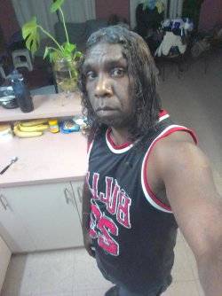 A Black man with long curly hair takes a selfie in a kitchen, wearing a black and red basketball jersey.