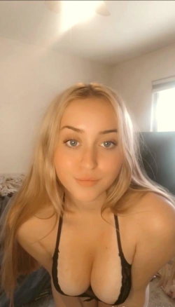 Blonde woman with blue eyes in a black lace bra, taking a close-up selfie in a softly lit bedroom.