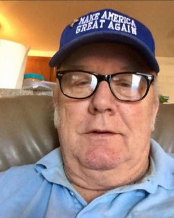 An older man with glasses and a blue cap sits on a couch wearing a light blue collared shirt.