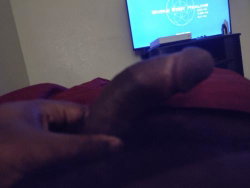 Close-up of a dark-skinned hand holding an erect penis, with a TV screen visible in the background.