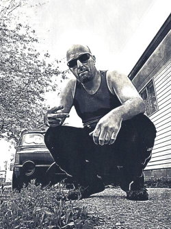 Bald man with sunglasses crouching outdoors, wearing a tank top and dark pants, near a vehicle and house.