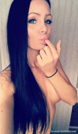 A woman with long dark hair and makeup poses topless, touching her lips with one hand in a close-up selfie.