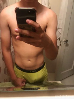 Shirtless man with toned torso taking mirror selfie in bright yellow underwear, holding phone in bathroom.