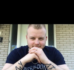A man with short blond hair and a beard, wearing a dark blue t-shirt, sits outdoors in front of a brick house with his
