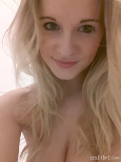 Blonde woman with wavy hair, bare shoulders, smiling softly in a close-up selfie indoors.