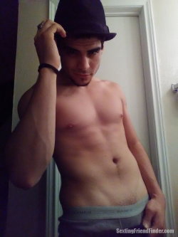 Shirtless man with defined abs wearing a fedora and gray underwear, standing indoors near a door.