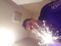 A man in a purple hoodie holds a sparkler indoors, smiling at the camera with bright sparks illuminating his face.