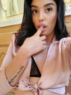 Brunette woman with dark wavy hair and a nose ring poses in a bathroom, wearing a pink robe over a black lace bra,