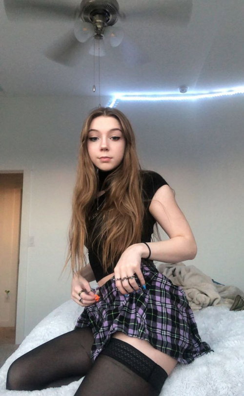 A young woman with long brown hair sits on a bed in a plaid skirt, black top, and thigh-high stockings, adjusting her