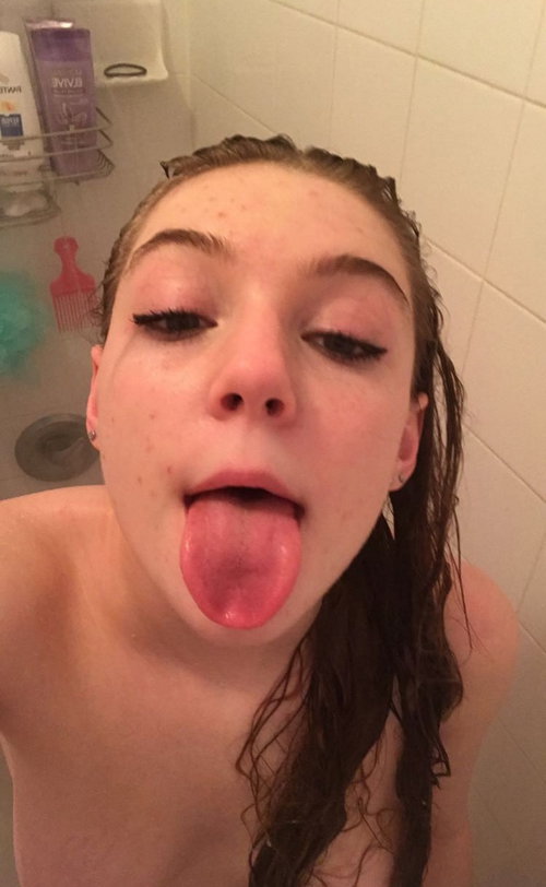 A young woman with wet dark hair sticks her tongue out in a bathroom selfie, bare shoulders visible.