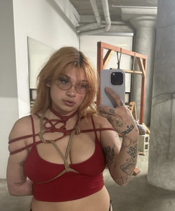 A woman with blonde hair and glasses takes a mirror selfie in a red strappy top, showcasing tattoos on her arm and hand