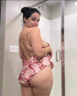 Curvy woman with dark hair in a bun, wearing a pink patterned robe, standing in a bathroom near a shower, looking over