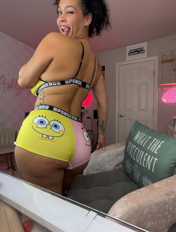 Curvy woman in SpongeBob-themed lingerie poses playfully in a pink room with neon signs and a mirror selfie setup.