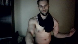 Shirtless muscular man with short beard and dark hair, flexing arm in dimly lit room, black shirt draped over shoulders.