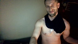 Shirtless man with short beard and dark hair, wearing a black neck wrap, smiling in a dimly lit indoor setting.