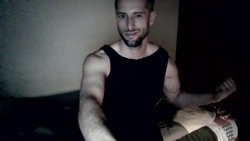 A muscular man with a beard wearing a black tank top, sitting indoors in dim lighting.