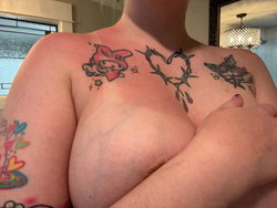 Topless woman with colorful chest tattoos, covering breasts with hand, indoors near window and chandelier.
