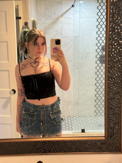 A young woman with colorful hair takes a mirror selfie in a black lace crop top and denim mini skirt, showing visible
