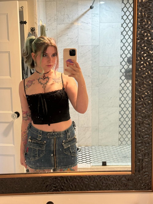 A young woman with colorful hair takes a mirror selfie in a black lace crop top and denim mini skirt, showing visible