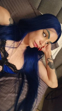 Woman with long blue hair and tattoos lies on a couch wearing a black lace top.