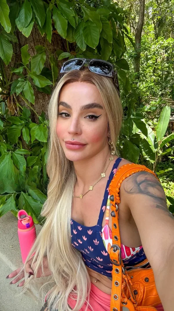 Slim blonde woman in blue crop top takes selfie outdoors amid lush green foliage.