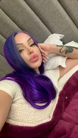 Woman with long purple hair lying on a bed in a white t-shirt taking a selfie.