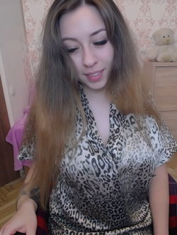 A woman with long ombre hair wears a leopard-print robe while sitting indoors, smiling softly in a cozy room with