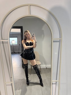 Woman with long hair in a black corset and pleated skirt poses in front of a mirror.